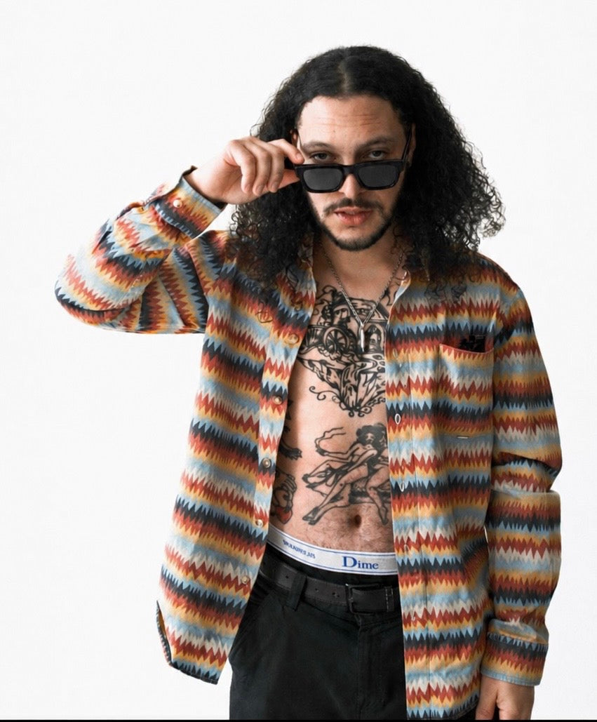 A man with long curly hair and a beard wearing a colorful zigzag knit cardigan, sunglasses, and a tattooed torso, posing against a plain white background.