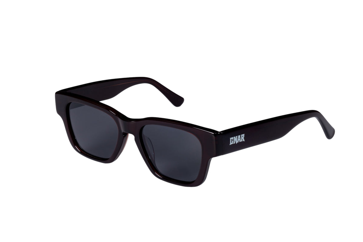 Front three quarter view of Gnar optics Disaster sunglasses with dark acetate burgundy rectangular frames, dark polarized lenses and a white GNAR logo on arms.